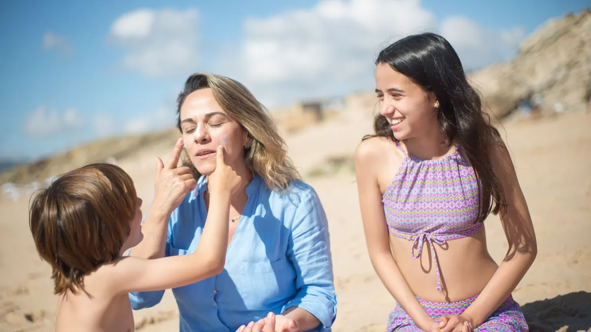 Menino passando protetor solar no rosto da mãe na praia, ao lado da irmã