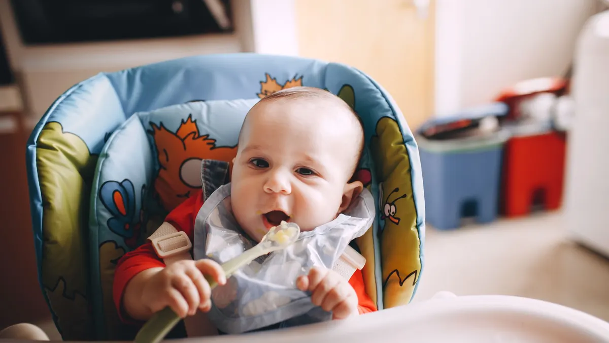 Bebê na cadeirinha de alimentação comendo papinha com a colher
