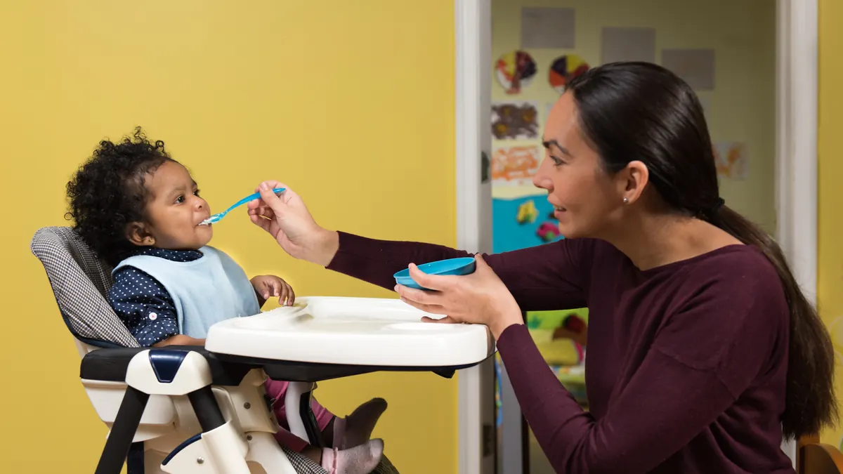 Mulher sentada diante de um bebê na cadeirinha de alimentação, oferecendo comida de colher