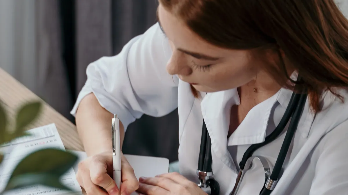 Médica de jaleco e estetoscópio escrevendo uma receita no consultório