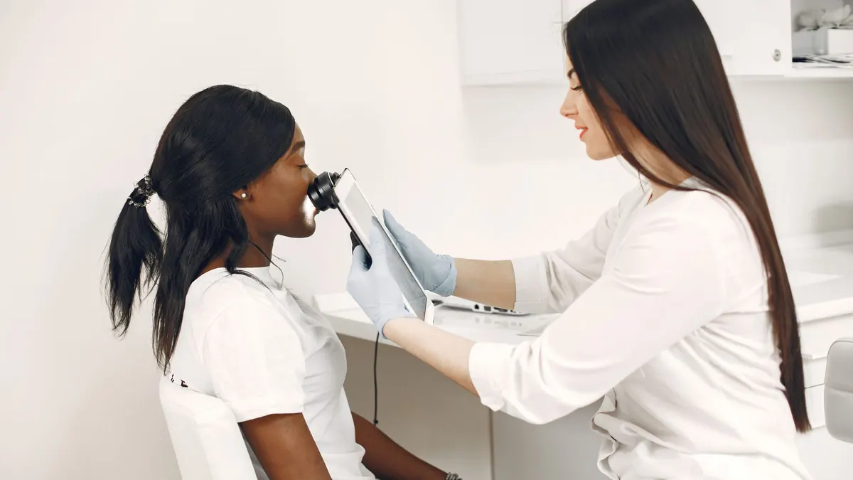 Dermatologista examinando a pele do rosto de uma mulher negra com um aparelho de aumento ligado a um tablet