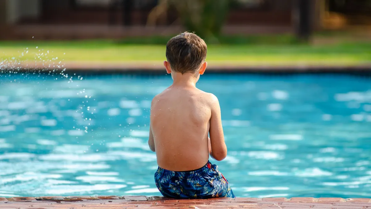 Menino de sunga estampada sentado na borda de uma piscina azul, de costas, com respingos de água no ar