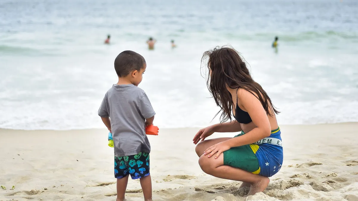 Mãe agachada na areia conversando com o filho pequeno na beira do mar, na praia de Copacabana, no Rio de Janeiro