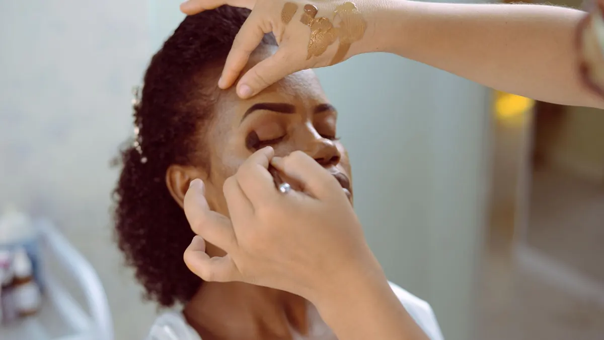 Maquiadora aplicando sombra nas pálpebras de uma mulher de cabelo cacheado, de olhos fechados