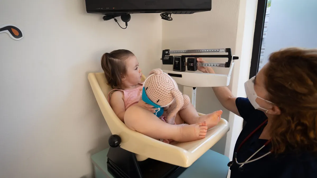 Menina pequena sentada na balança do consultório enquanto a pediatra confere o peso