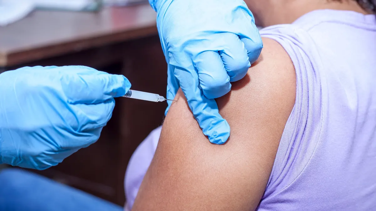 Profissional de luvas aplicando uma injeção no braço de uma pessoa de blusa lilás