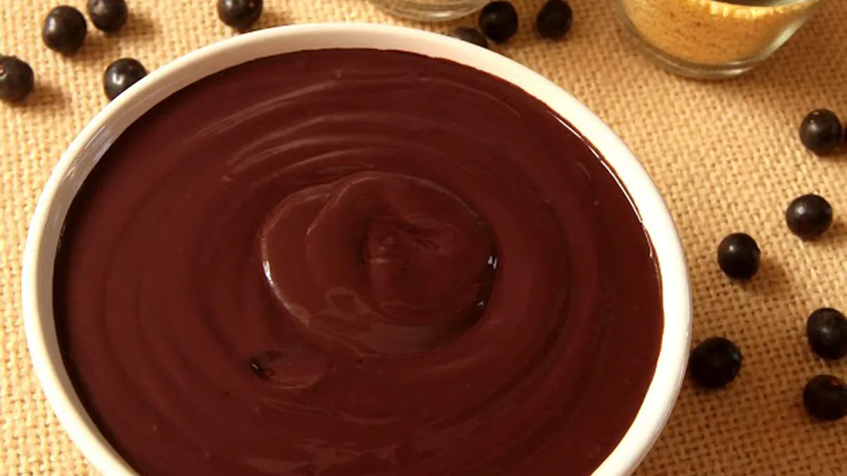 Açaí na tigela, de perto, com grãos de açaí espalhados em volta e potinhos de farinha de tapioca e granola ao fundo