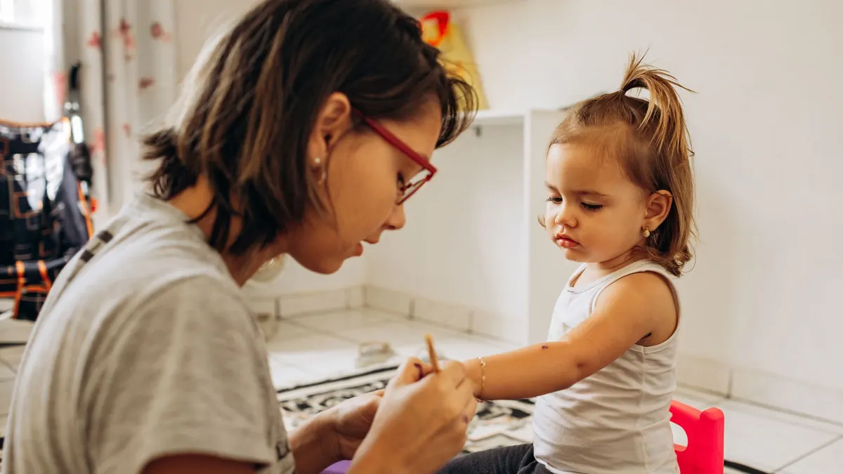 Mulher cuidando do braço de uma menina pequena, sentadas no chão de casa