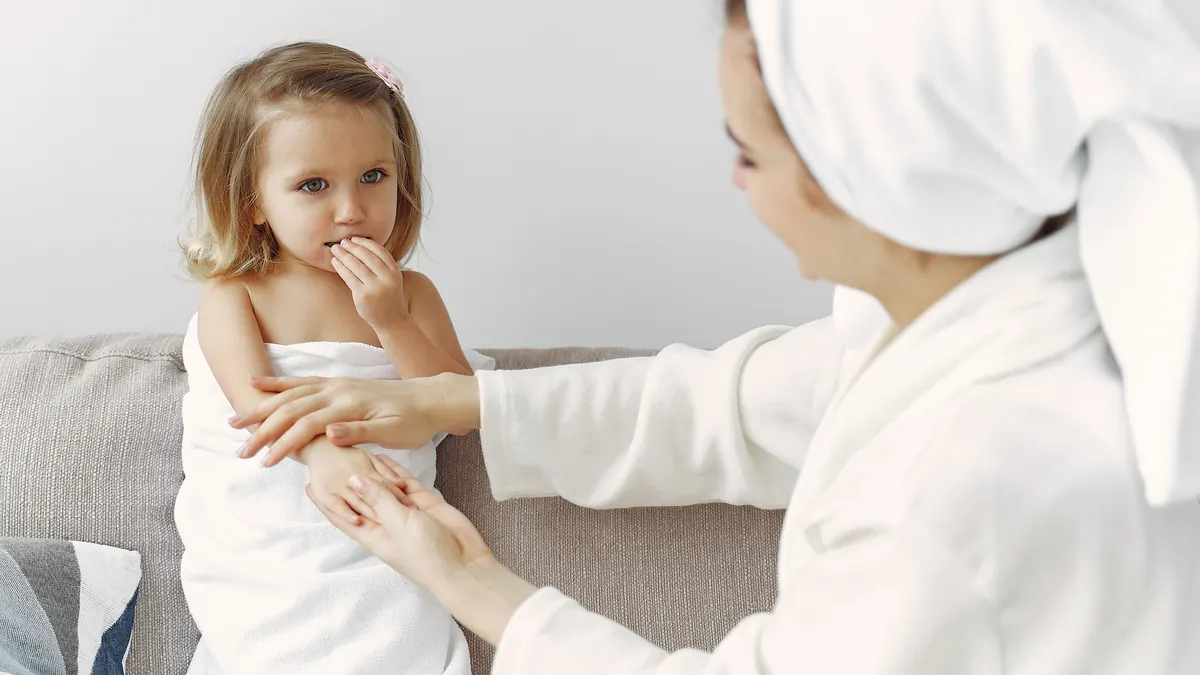 Mãe passando creme no braço da filha pequena enrolada na toalha, depois do banho