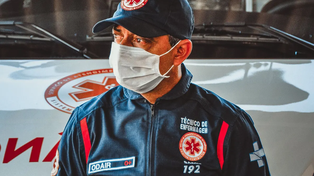 Técnico de enfermagem do SAMU 192, de boné e máscara, em frente a uma ambulância